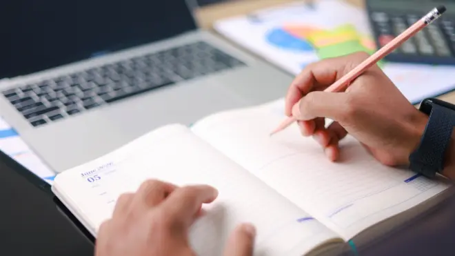 Business professional writing notes in planner beside laptop and charts. Concept of productivity, scheduling, planning, strategy, and office workflow.